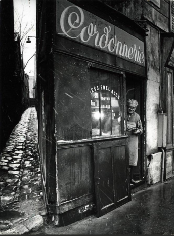 Black and white photo of a small shop in a European city, maybe Paris. The owner stands in the doorway. The photo has some streaks that might be from falling rain or falling snow.