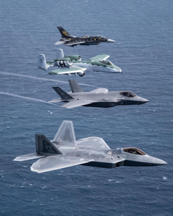 Color photo of four US Air Force fighter or attack jets flying in formation over open water. From front to rear we see an F-22, an F-35, an A-10, and an F-16. The paint schemes on the last two jets look unusual as if they're painted for a special occasion.