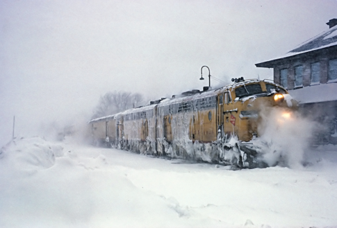 Color photo of a Milwaukee Road passenger train with two diesels up front, at the depot in Calumet, Michigan. The days seems to be really frigid cold -- the locomotives are caked in ice and snow.