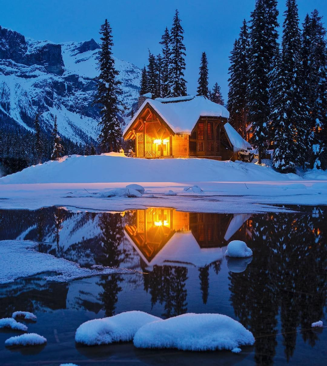 Nighttime color photo of a 2 story house in a snowy and mountainous wilderness with a lake in the foreground. The house is lit from inside with a beautiful golden glow that reflects on the water of the lake.